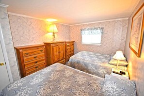 Ein Schlafzimmer mit zwei Betten, Holzkommoden, einem Fenster mit Vorhang und einem Nachttisch mit Lampe.