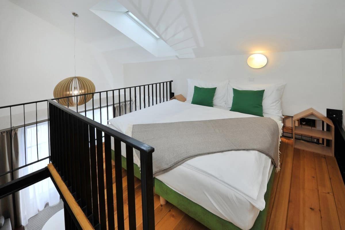 Un dormitorio con tragaluz, cama con ropa de cama verde y blanca, lámpara colgante y una pequeña estructura de madera.