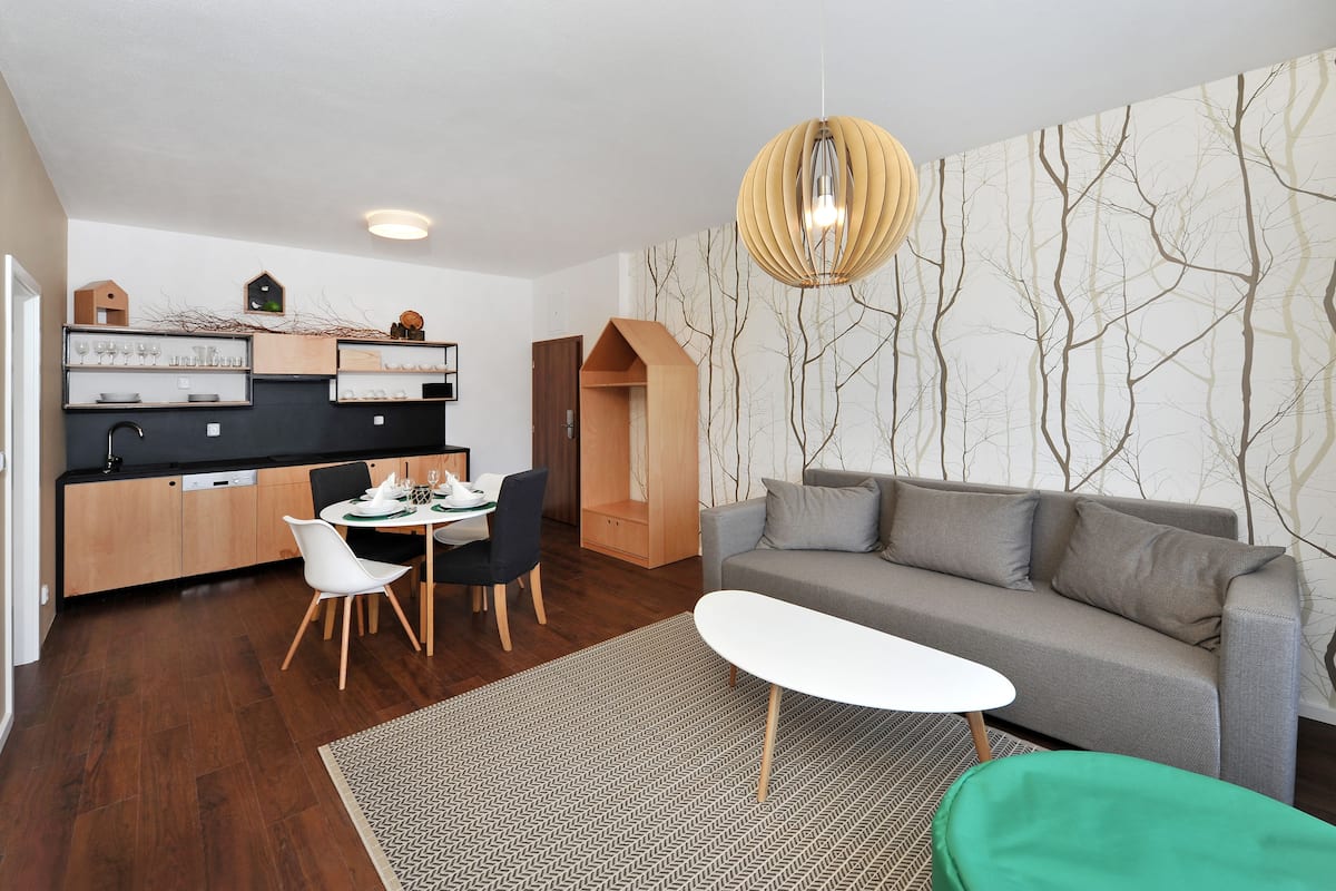 Una sala de estar moderna con un sofá gris, una mesa de comedor con sillas, una mesa de centro blanca y un reposapiés verde. Se observa un área de cocina con gabinetes de madera y un colgante.