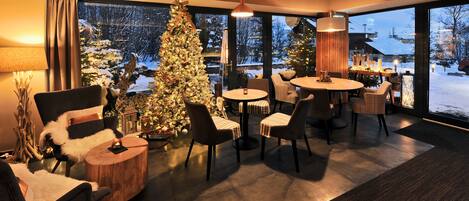 A modern living room with a large Christmas tree, comfortable seating, and a view of a snowy landscape through large windows.
