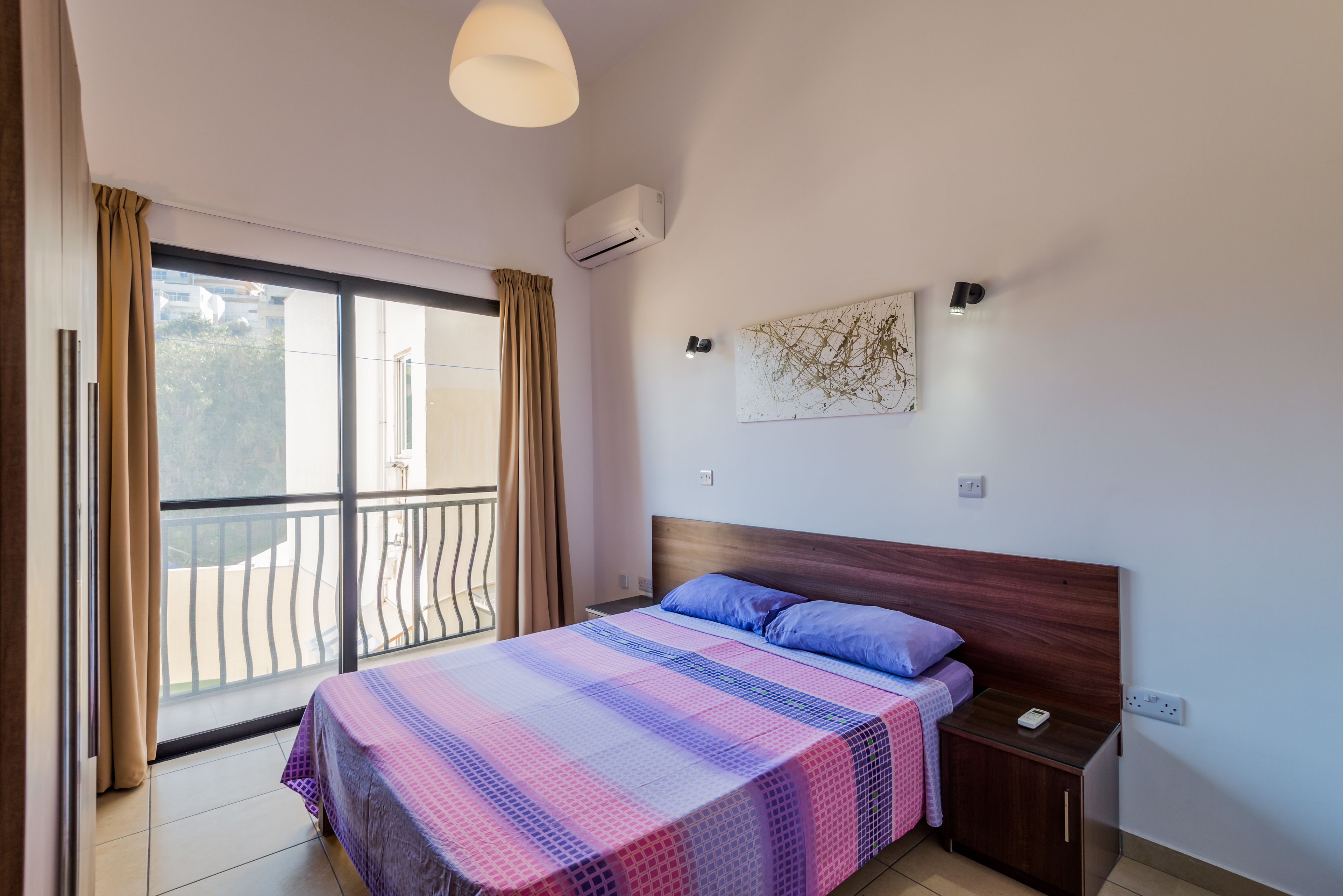 Un dormitorio con cama, mesita de noche, aire acondicionado instalado en la pared, ventana con cortinas y balcón.