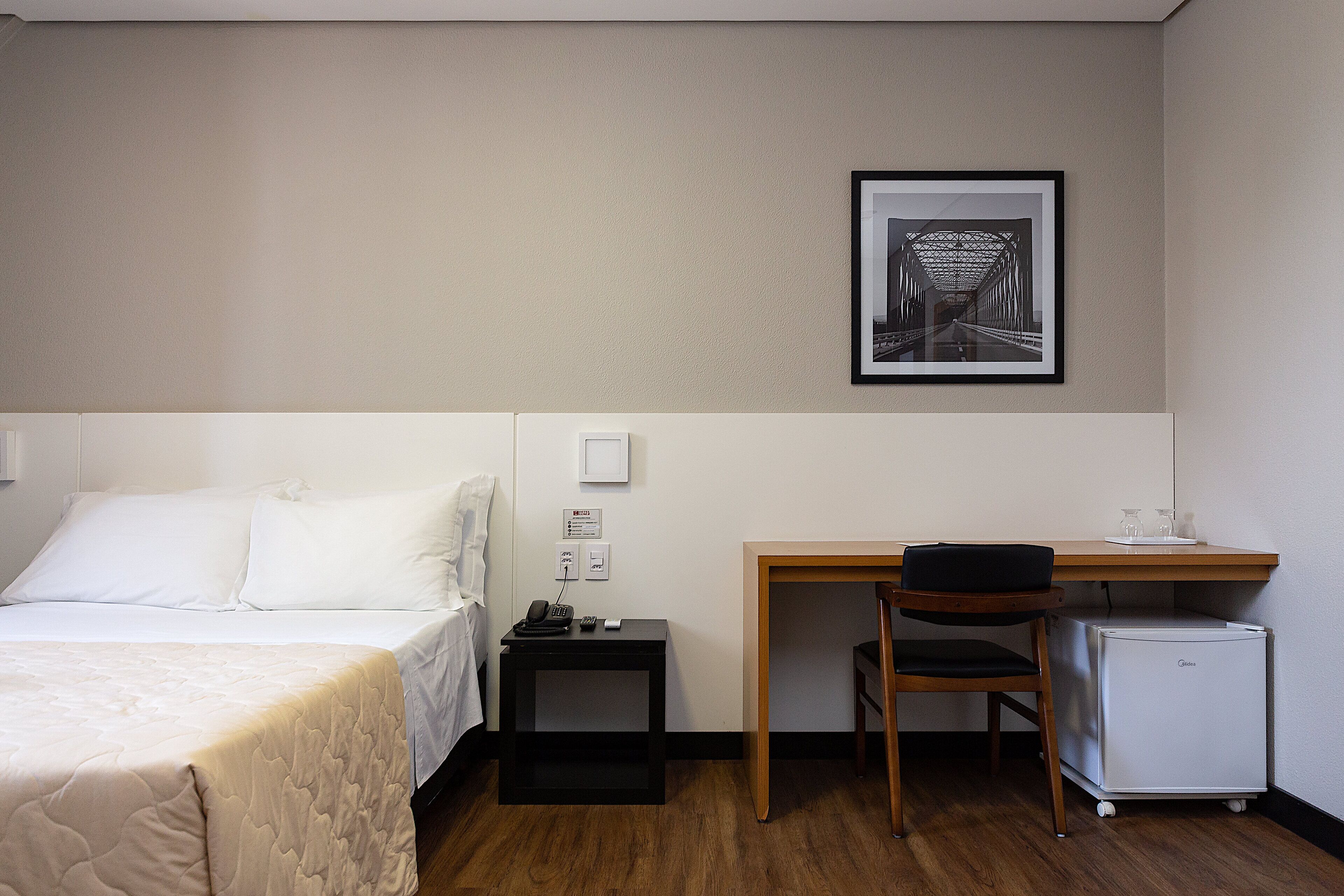 Ein Hotelzimmer mit Bett, Schreibtisch, Stuhl, kleinem Kühlschrank und einem gerahmten Bild an der Wand.