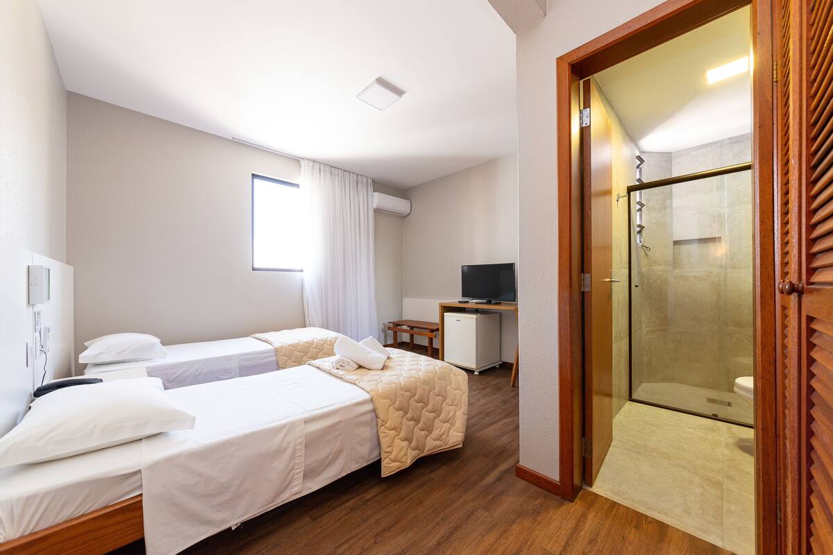 A hotel room with two beds, a desk with a computer, a shower area, and a window with curtains.