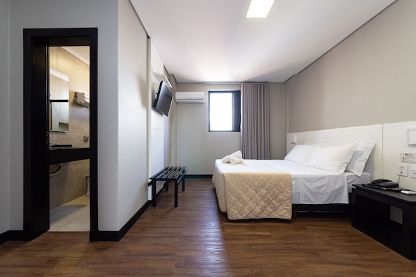 A hotel room with a bed, a bench, a TV, an air conditioner, and a window with curtains.