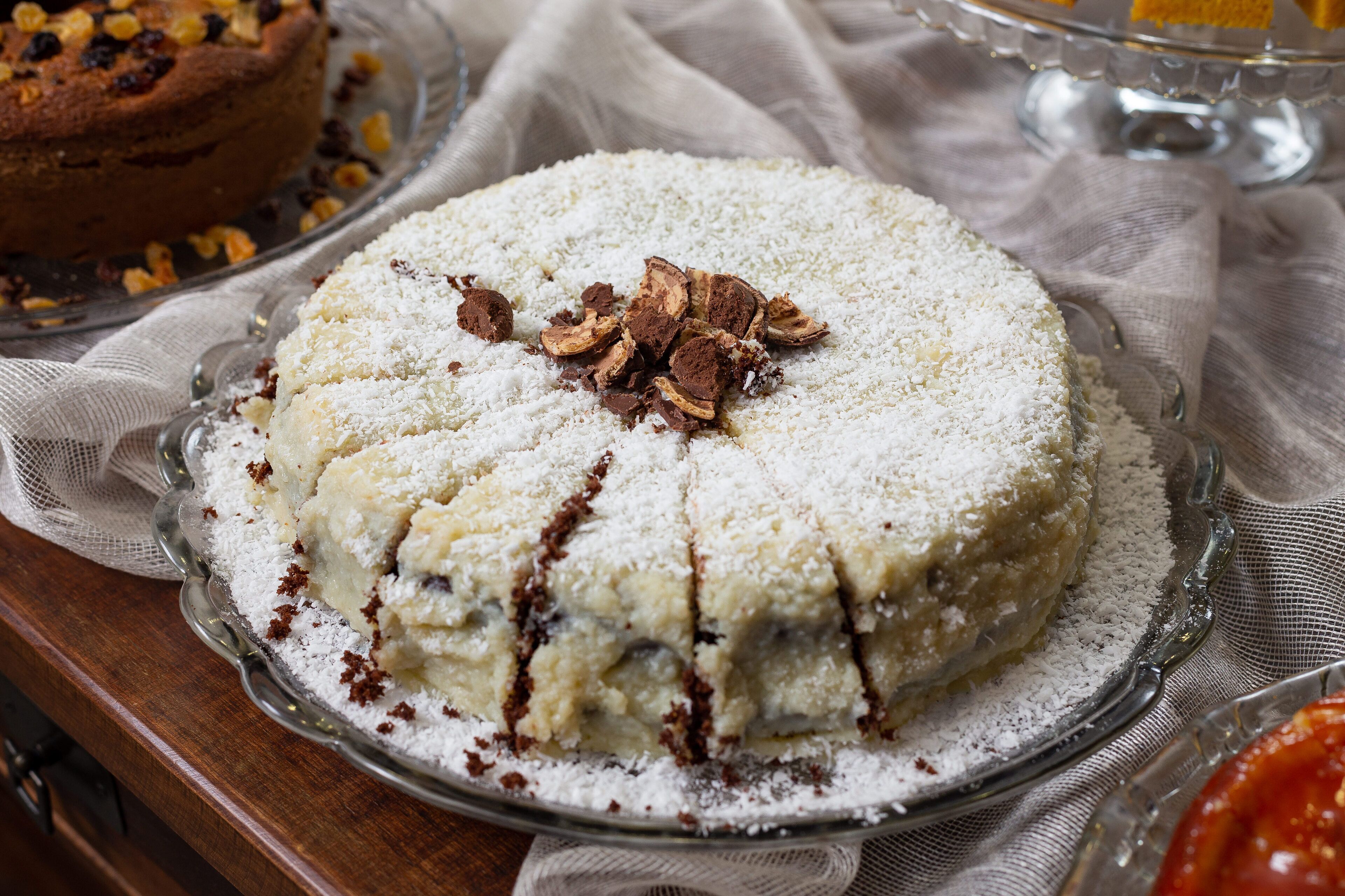 Un pastel redondo con glaseado blanco y virutas de chocolate sobre un expositor de vidrio.