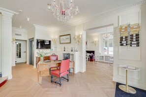 A spacious room with a wooden floor, a desk with a chair, a television, and a chandelier. - ACANTHE HOTEL (Bordeaux)
