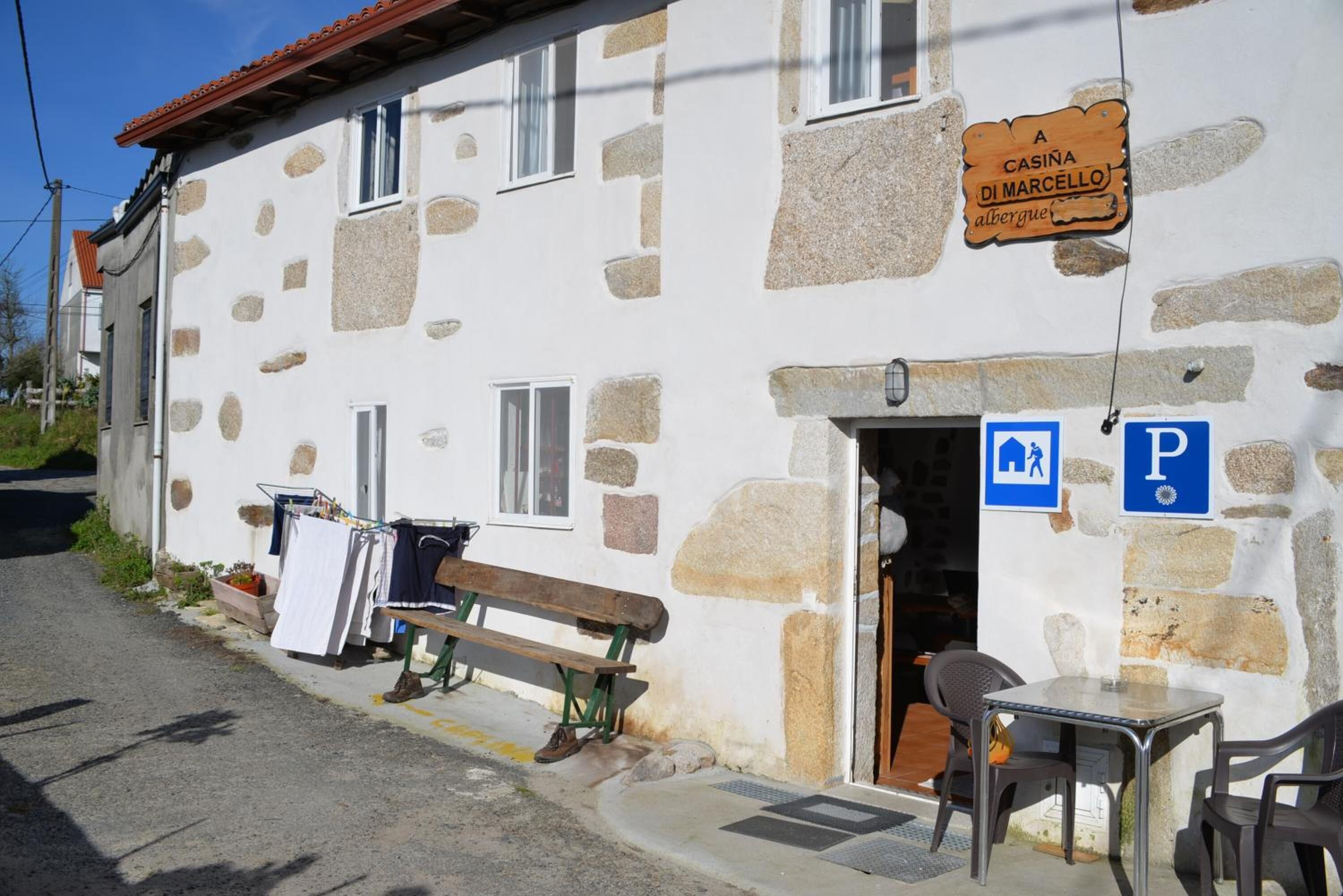 A stone building with a sign reading "CASINA DI MARCELLO". There is a bench outside, a table with chairs, and a parking sign.