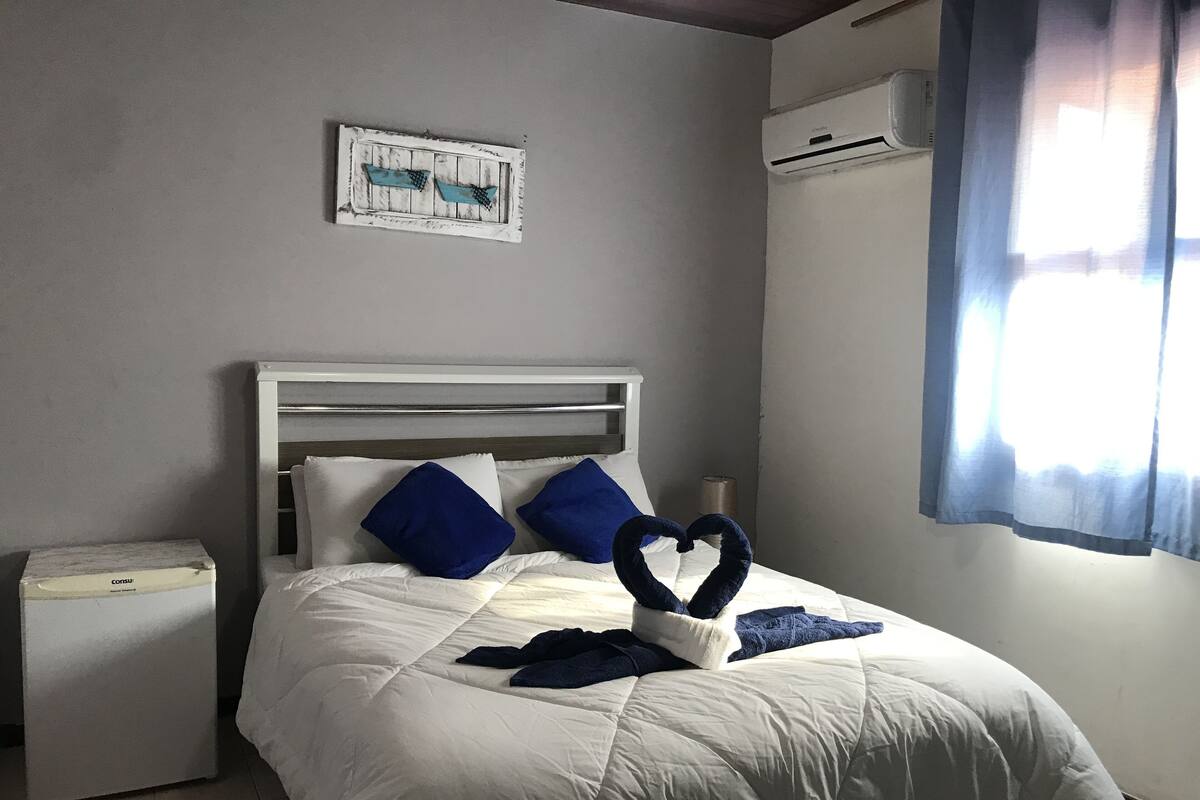 Una cama bien hecha con ropa de cama blanca y almohadas azules, una mesita de noche pequeña, un aire acondicionado de pared y una ventana con cortinas.