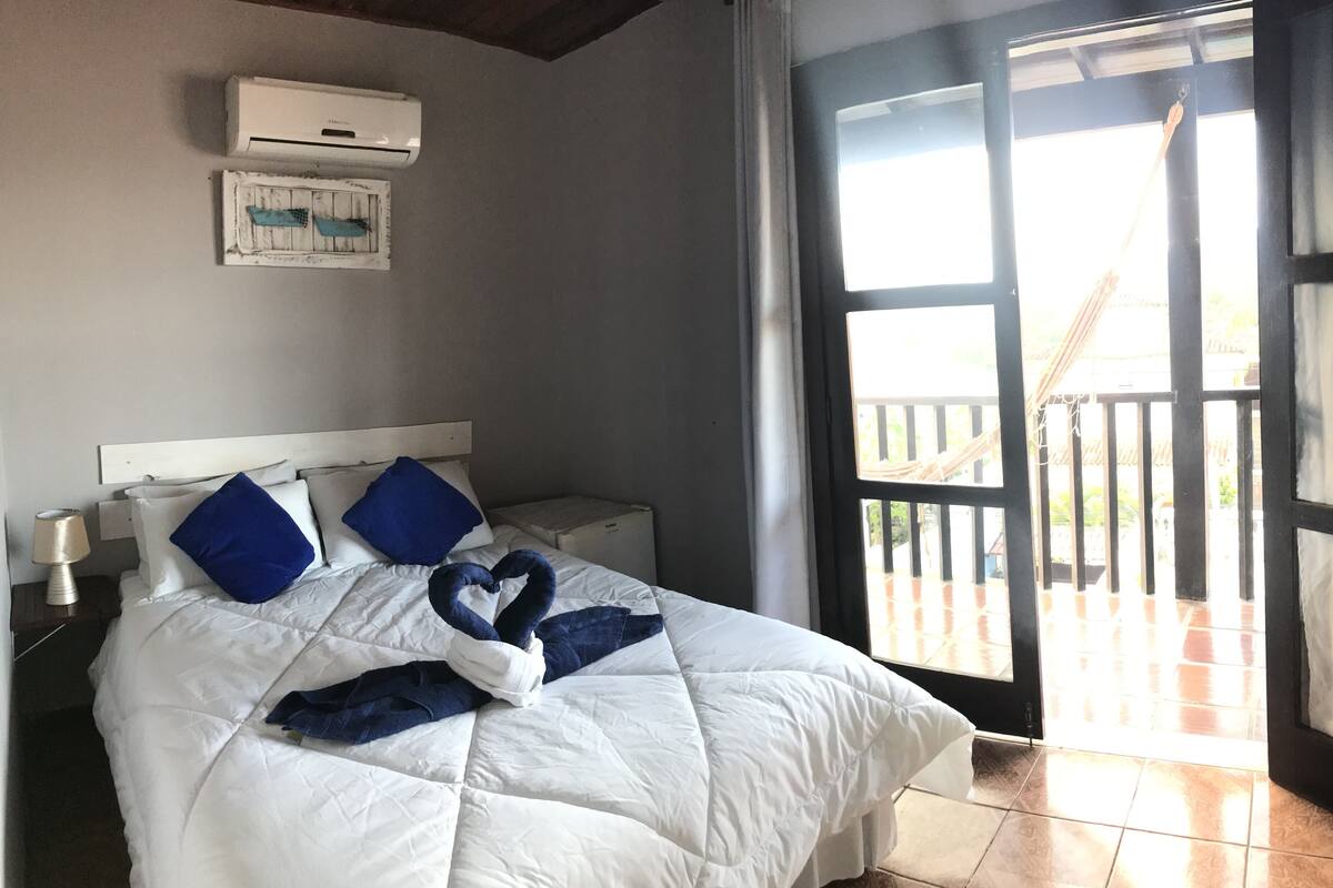Una habitación de hotel con cama, mesita de noche, aire acondicionado, ventana con vista a un balcón y puerta.