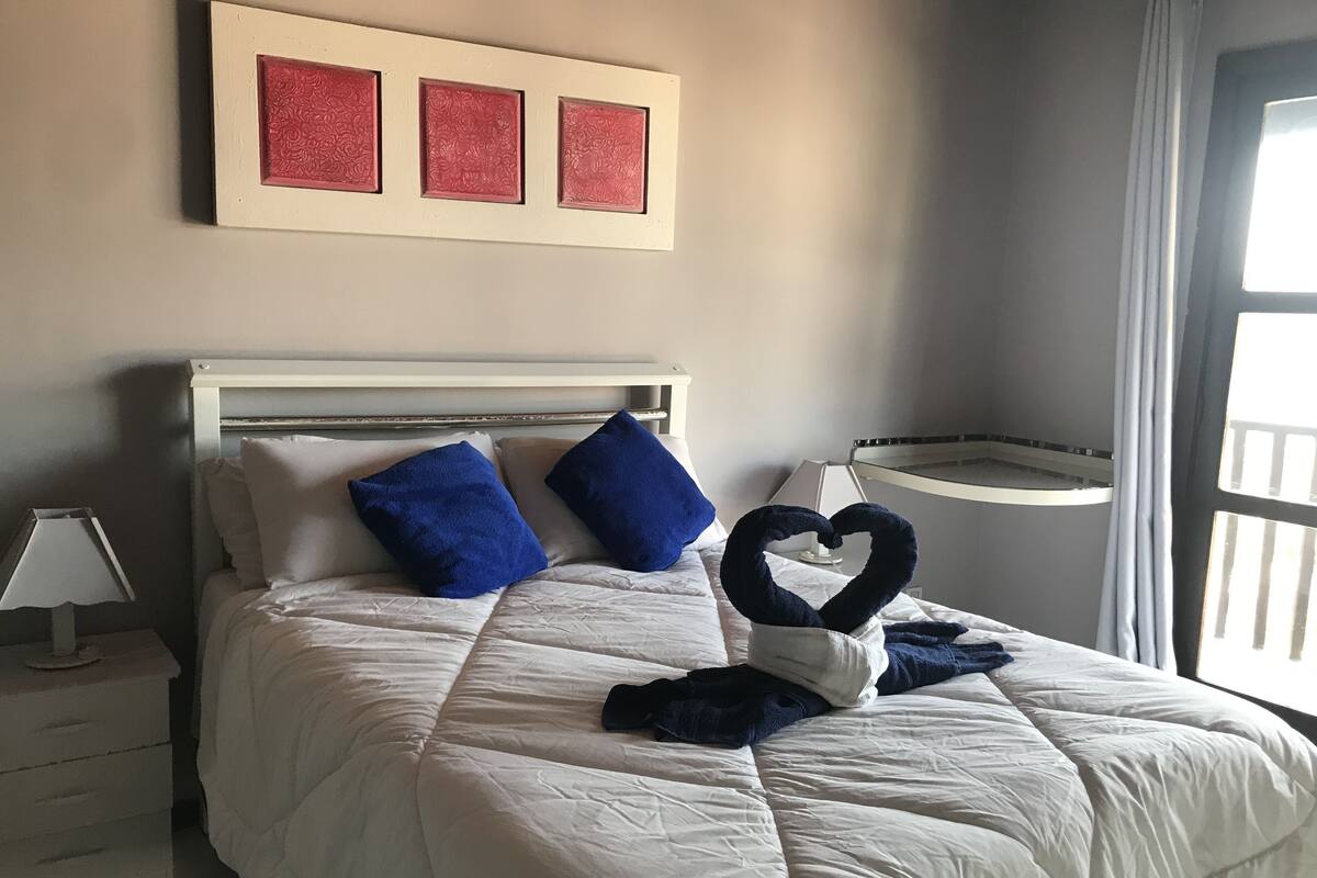 Una cama bien tendida con ropa de cama blanca y almohadas azules, un arreglo de toallas con forma de corazón y una mesita de noche con una lámpara.