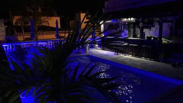 Uma área de piscina ao ar livre à noite, iluminada em azul, com um bar e um restaurante chamado 'Heineken'.