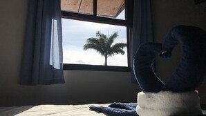 A bed with a blue and white towel shaped like a swan, a window with a view of a palm tree, and a curtain partially covering the window. - Pousada Azul dos Búzios (Búzios)