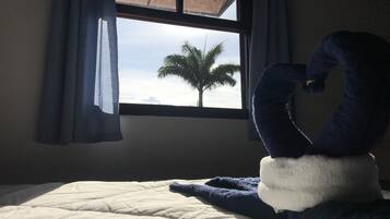 Uma cama com uma toalha azul e branca em formato de cisne, uma janela com vista para uma palmeira e uma cortina cobrindo parcialmente a janela.