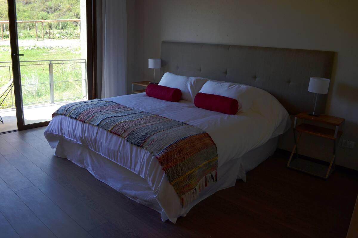 Una cama grande con cabecera, dos mesitas de noche con lámparas, una manta tejida de colores y vistas al verdor a través de una puerta de vidrio.