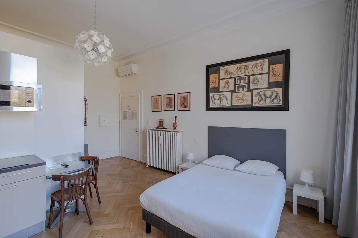 A modern hotel room with a bed, a desk, and a chair. There are framed pictures on the wall and a radiator.