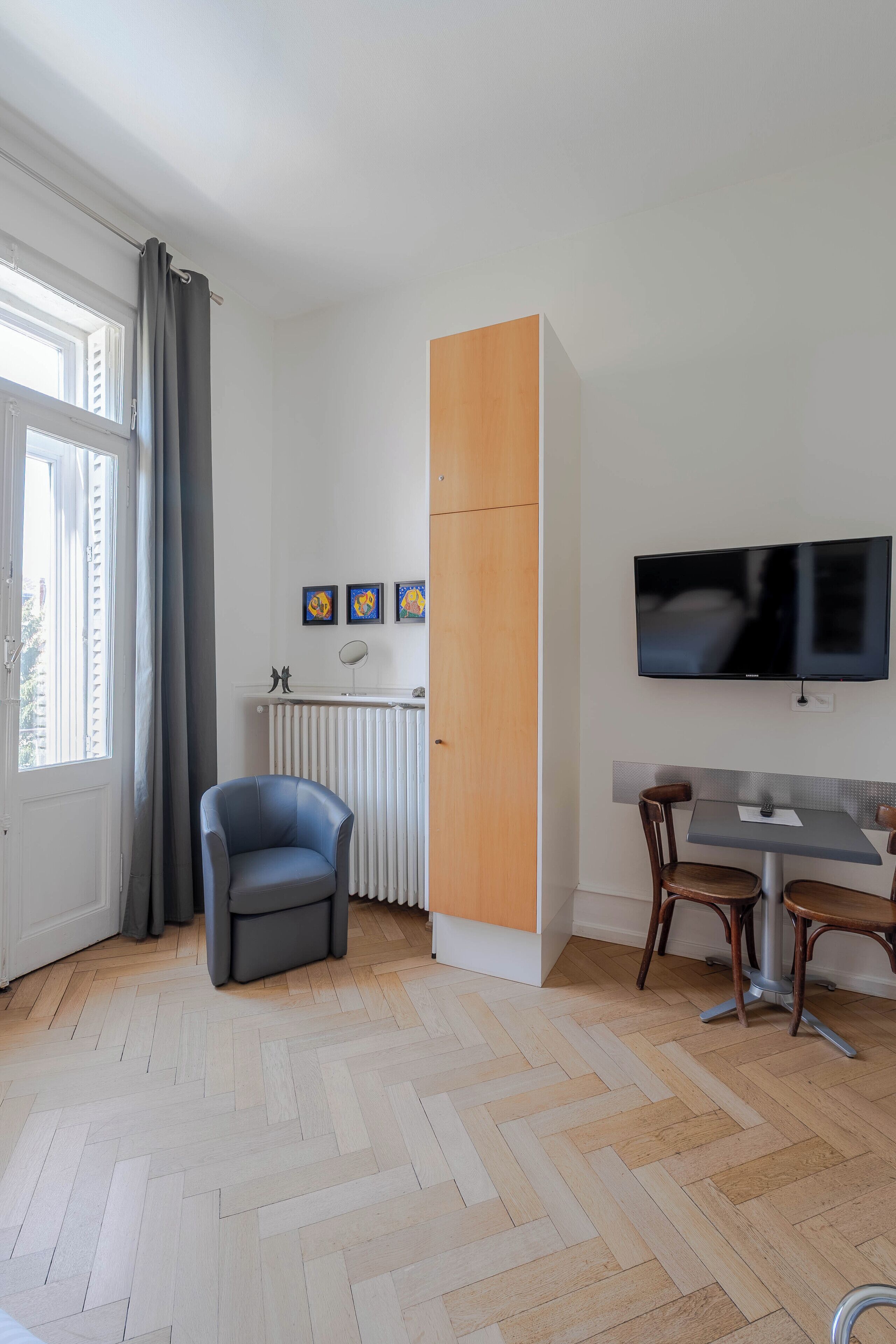 A modern living room with a herringbone floor, a grey armchair, a wooden cabinet, a TV mounted on the wall, and a dining area with a table and chairs.