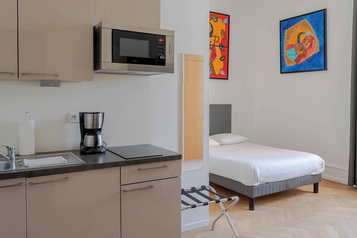 A compact room with a kitchenette, a bed, and two colorful paintings on the wall.