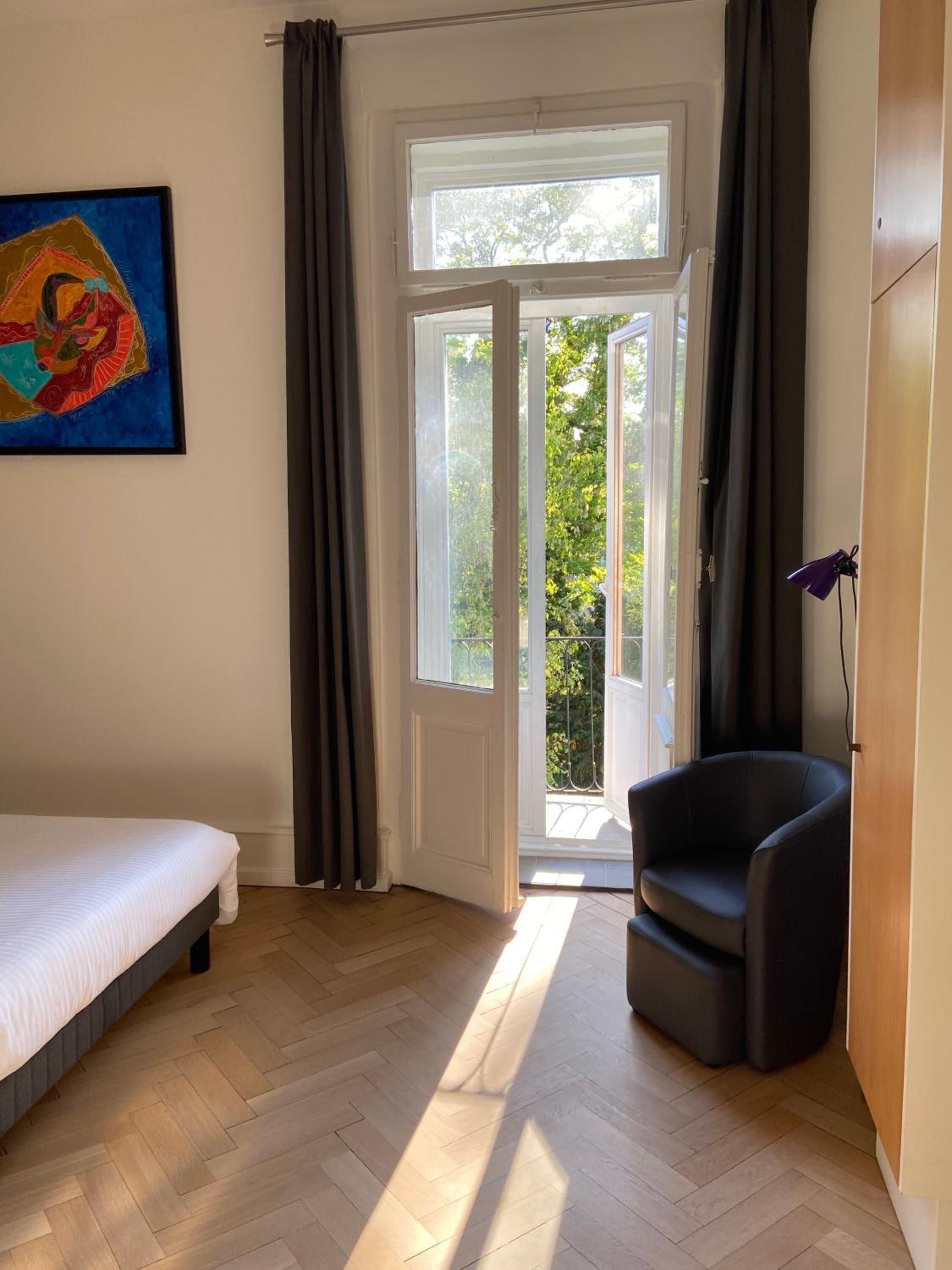 Ein Zimmer mit einem Bett, einem Sessel, einem Gemälde und Blick durch eine Glastür nach draußen.