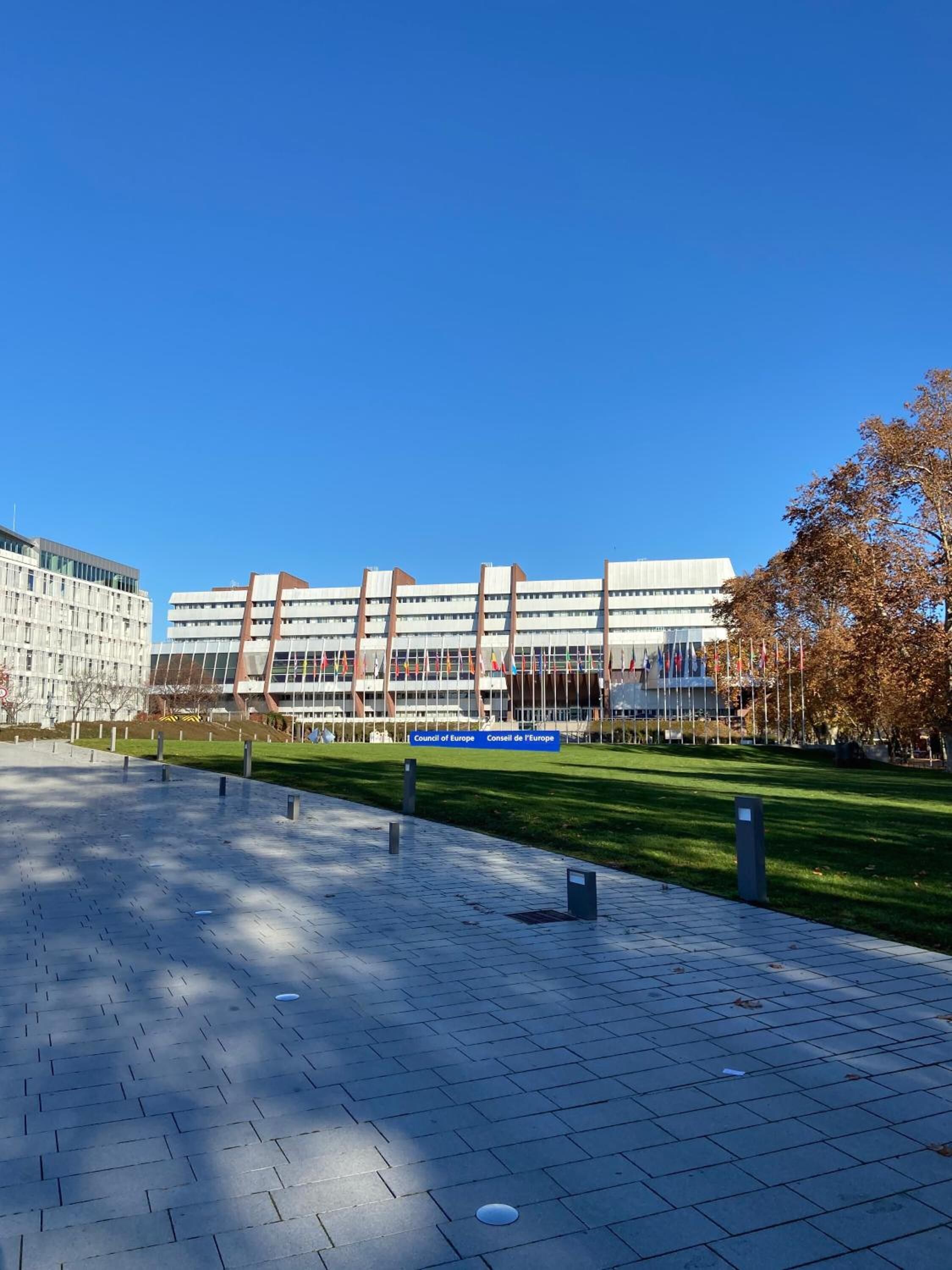 En modern byggnad med ett plattbelagt torg framför, där ett skylt med texten ”Council of Europe Consell de l’Europa” är placerat.