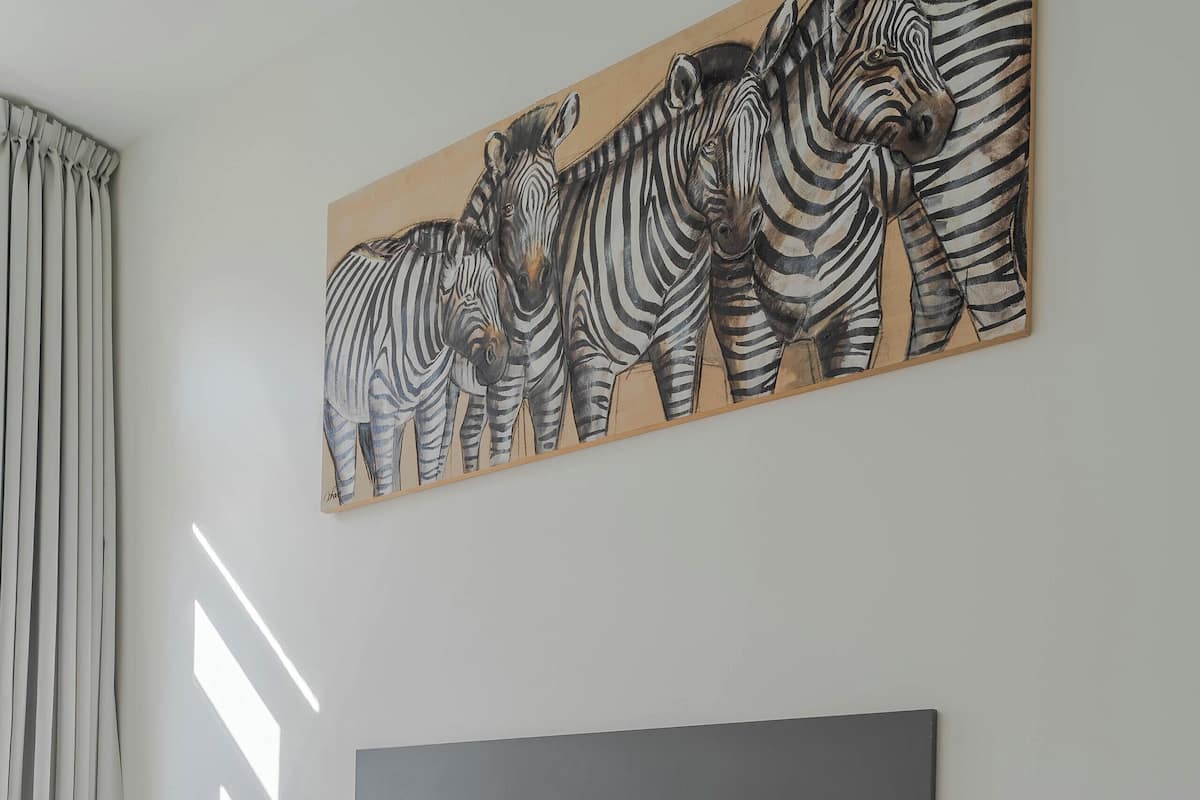 A modern bedroom with a grey headboard, white bedding, a bedside table with a lamp, and a zebra painting on the wall.