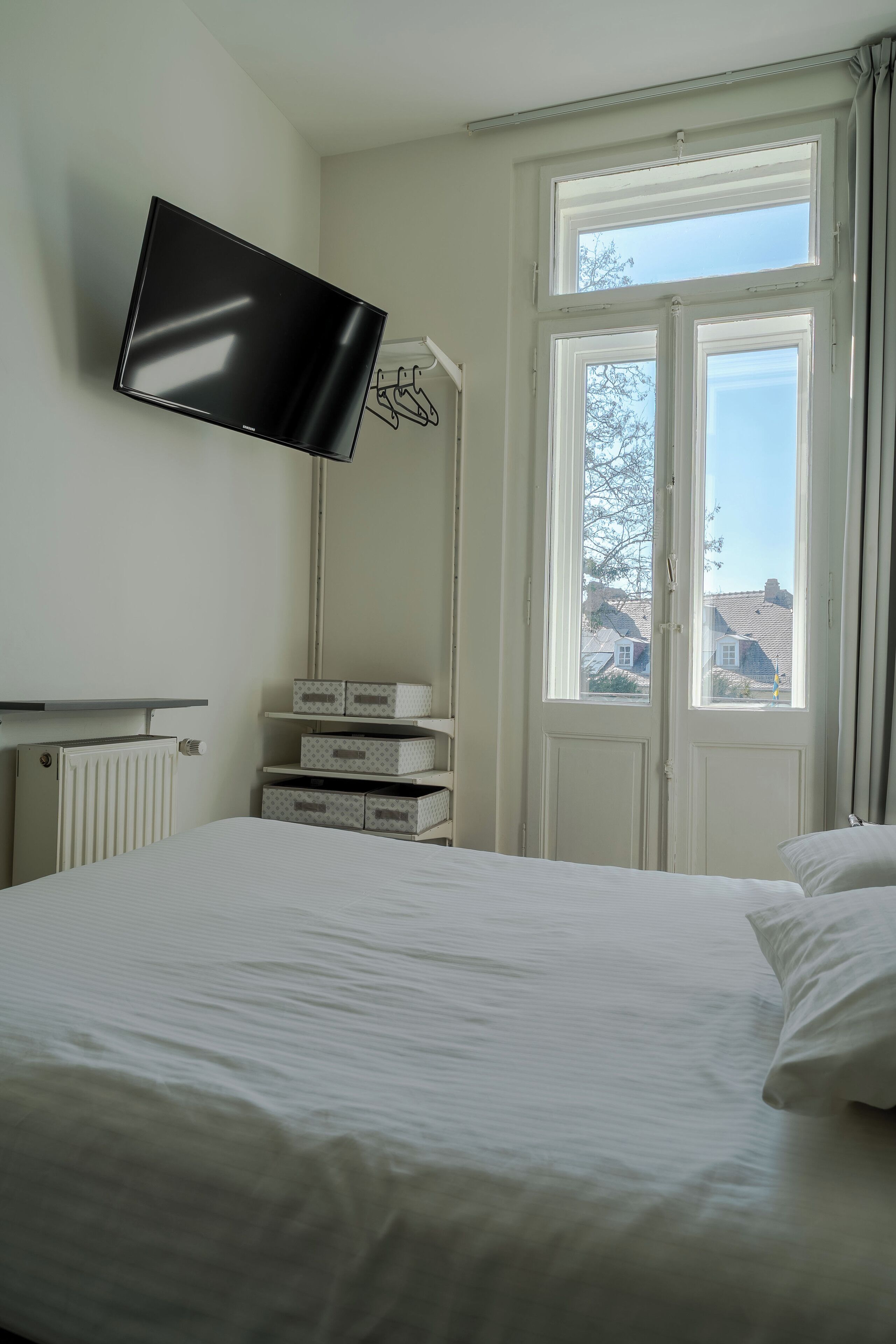 Ein Hotelzimmer mit Bett, Wandfernseher, Fenster mit Vorhängen und Aufbewahrungsboxen.
