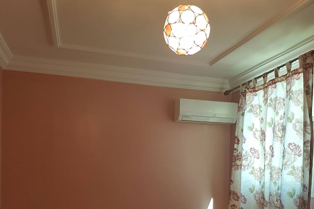 A bedroom with a bed, pillows, a patterned bedspread, an air conditioning unit, a nightstand, and a window with floral curtains.