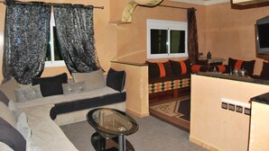En stue med sofa, sofabord i glas og et køkkenområde med bordplade og hårde hvidevarer.
