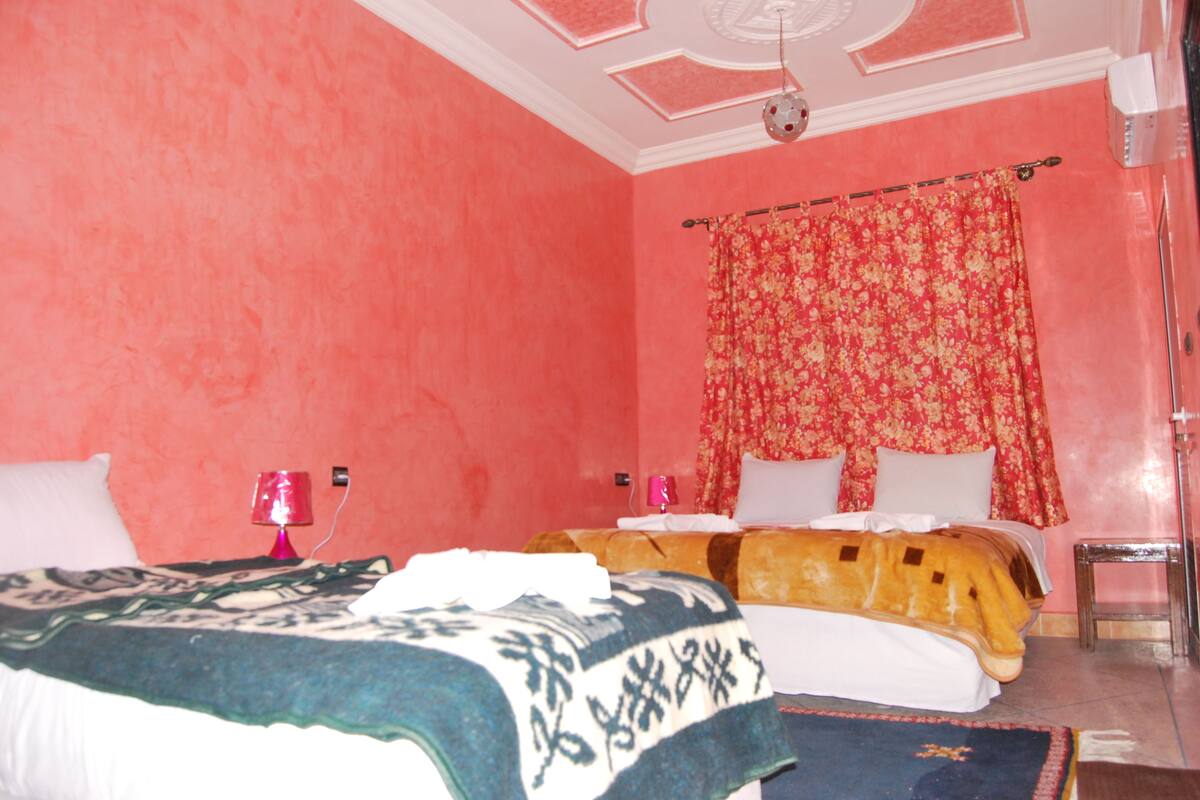 A room with pink walls, two beds with patterned bedding, a red curtain, and a ceiling fan.
