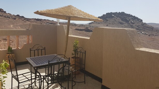En terrasse med et bord og stole, en stråparasol og udsigt til stenede omgivelser.