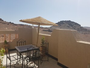 A terrace with a table and chairs, a thatched umbrella, and a view of rocky terrain. - Résidence Marwa (Tarmigt)