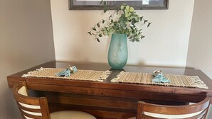A wooden dining table with two chairs, a vase with green foliage, and a framed picture above. - Beautiful 4th floor Gulf View Condo (Hudson)