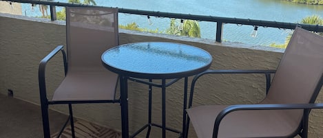 A balcony with a round table and two chairs, overlooking a body of water with trees and a distant shoreline.