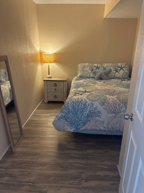 A bedroom with a bed featuring a coral pattern, a nightstand with a lamp, and a wooden floor.