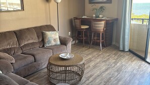 A living room with a brown sectional sofa, a round wicker coffee table, and a dining area with a small table and chairs. - Beautiful 4th floor Gulf View Condo (Hudson)