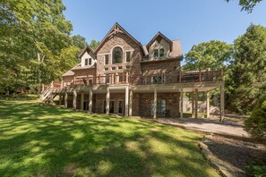 Une grande maison en pierre à deux étages, dotée d’un porche couvert et d’une terrasse entourée d’une clôture.