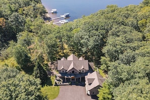 An aerial view of a large, multi-story house with a symmetrical facade, surrounded by dense forest and a dock with boats.