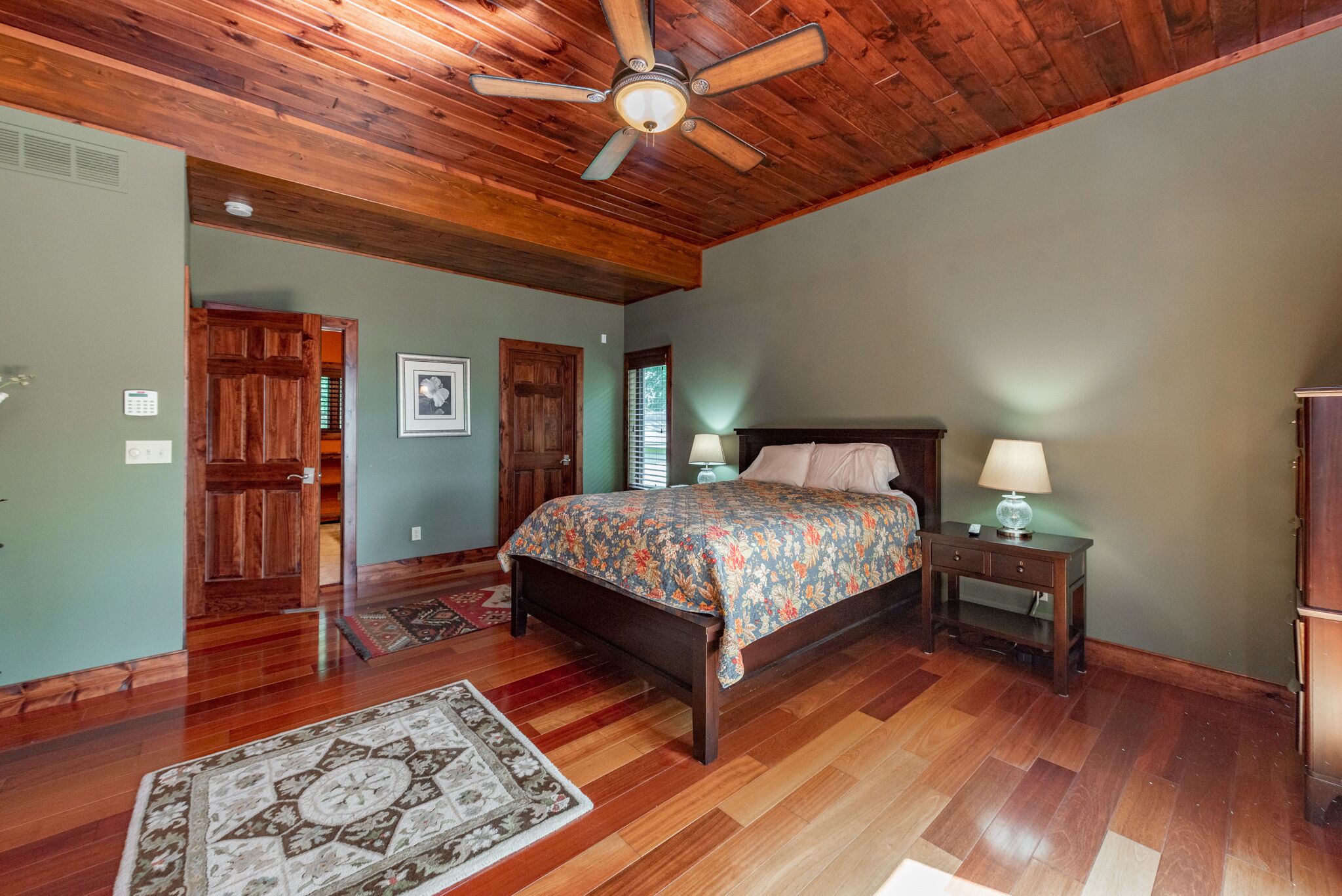 A bedroom with a wooden ceiling, a bed with a floral bedspread, two bedside tables with lamps, and a patterned rug on the floor.