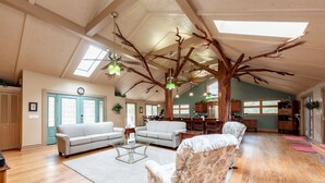 Ein geräumiges Wohnzimmer mit hoher Decke, Holzbalken und einer großen Baumskulptur. Zwei Sofas, ein Couchtisch und ein Teppich sind vorhanden.