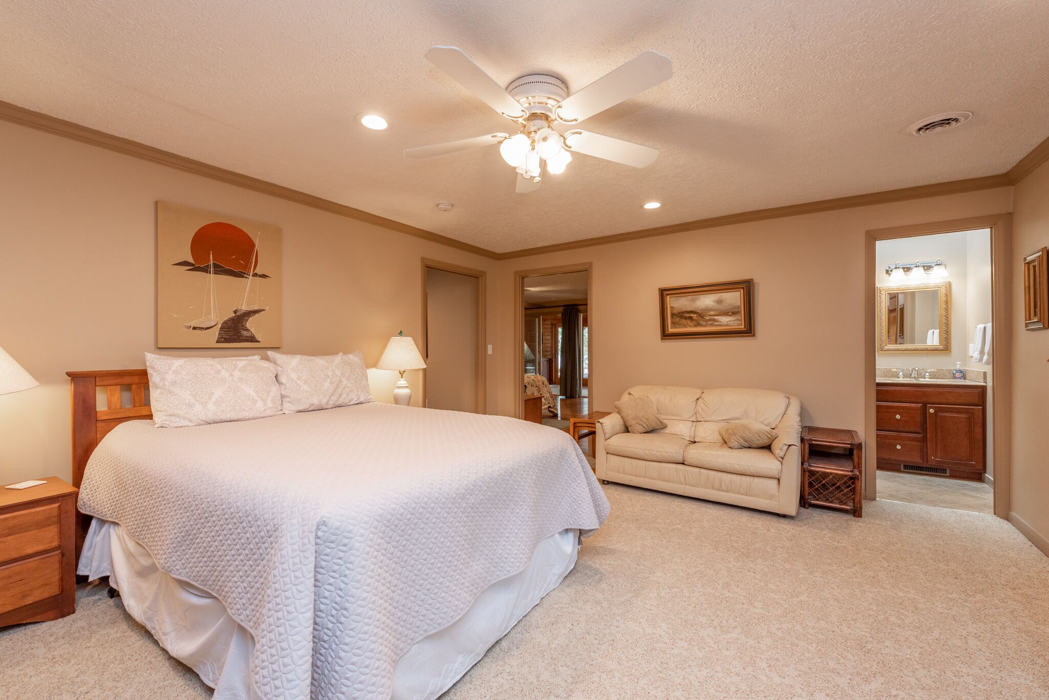 A bedroom with a large bed, a sofa, a ceiling fan, a nightstand, and a bathroom visible through an open doorway.