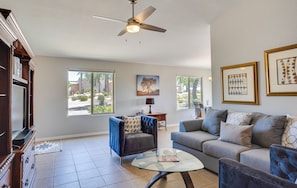 A spacious living room with a ceiling fan, large windows, a fireplace, and a view of the outdoors.