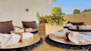 A dining area with a wooden table set for four, featuring black placemats and white napkins. There is a decorative plant with yellow flowers in a wooden planter. - 3BR w pool, golf community, dog friendly (Maricopa)