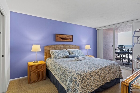 A bedroom with a bed, bedside lamps, a wooden headboard, a patterned bedspread, and a view of the balcony with outdoor furniture.