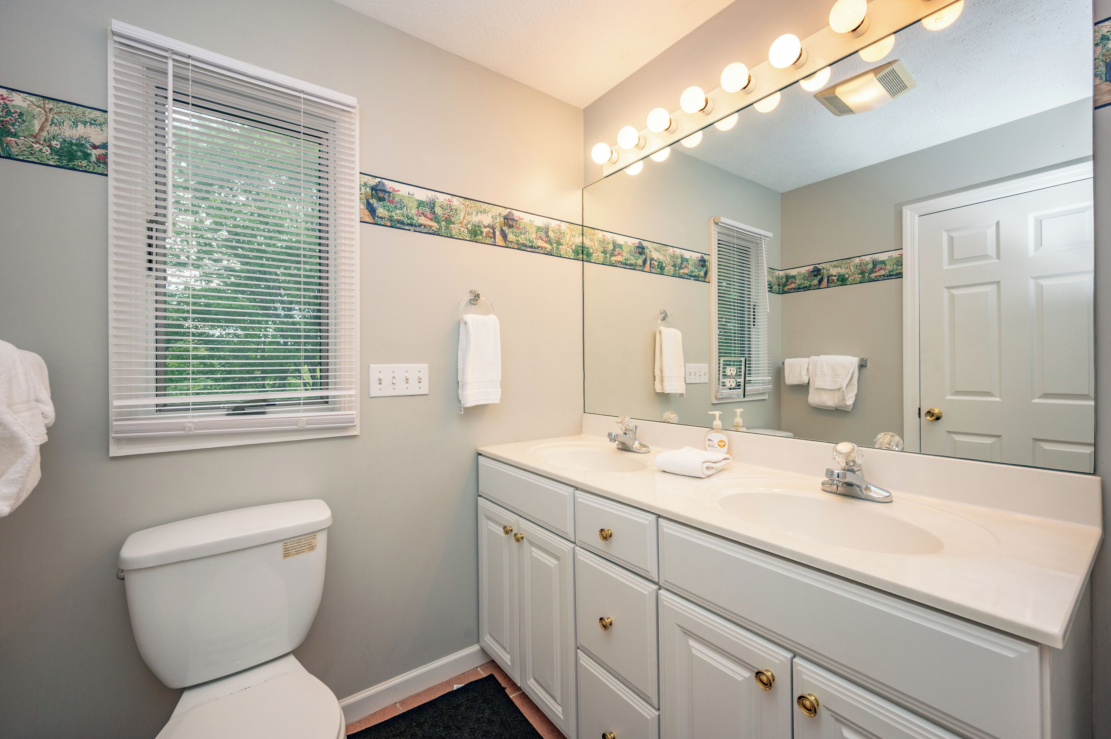 Une salle de bain comprenant des toilettes, un lavabo avec un miroir au-dessus et une fenêtre avec des stores.