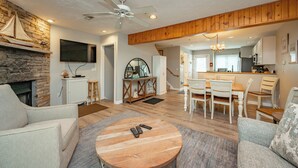 A spacious living room with a stone fireplace, a dining area with white chairs, and a kitchen in the background.