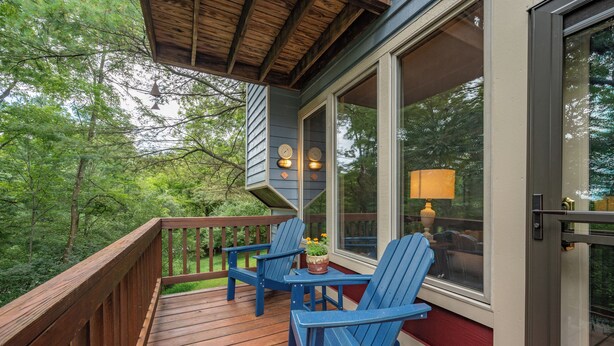 Una terraza de madera con sillas Adirondack azules y una mesa, con vistas a una zona boscosa.