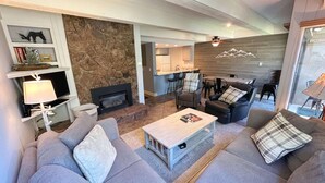 A modern living room with a stone accent wall, a fireplace, a dining area with a bar, and a kitchen in the background. - Super Location Between Lionshead and Vail Village - Outdoor Pool and Hot Tub (Vail)