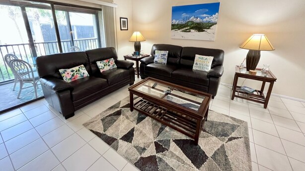 A living room with a sofa, coffee table, and two lamps. There is a picture on the wall and a chair outside the window. - Relax and enjoy your tropical getaway in this lovely Silver-rated condo! (Saint Augustine)