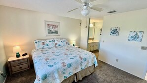 A bedroom with a bed, nightstand, lamp, ceiling fan, and artwork on the walls. - Relax and enjoy your tropical getaway in this lovely Silver-rated condo! (Saint Augustine)