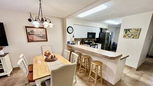 A modern kitchen with white cabinets, a microwave, and a refrigerator. There is a dining area with a wooden table and chairs, and a small bar counter with stools. - Enjoy family time in this Gold-rated condo with quick access to the beach! (Saint Augustine)