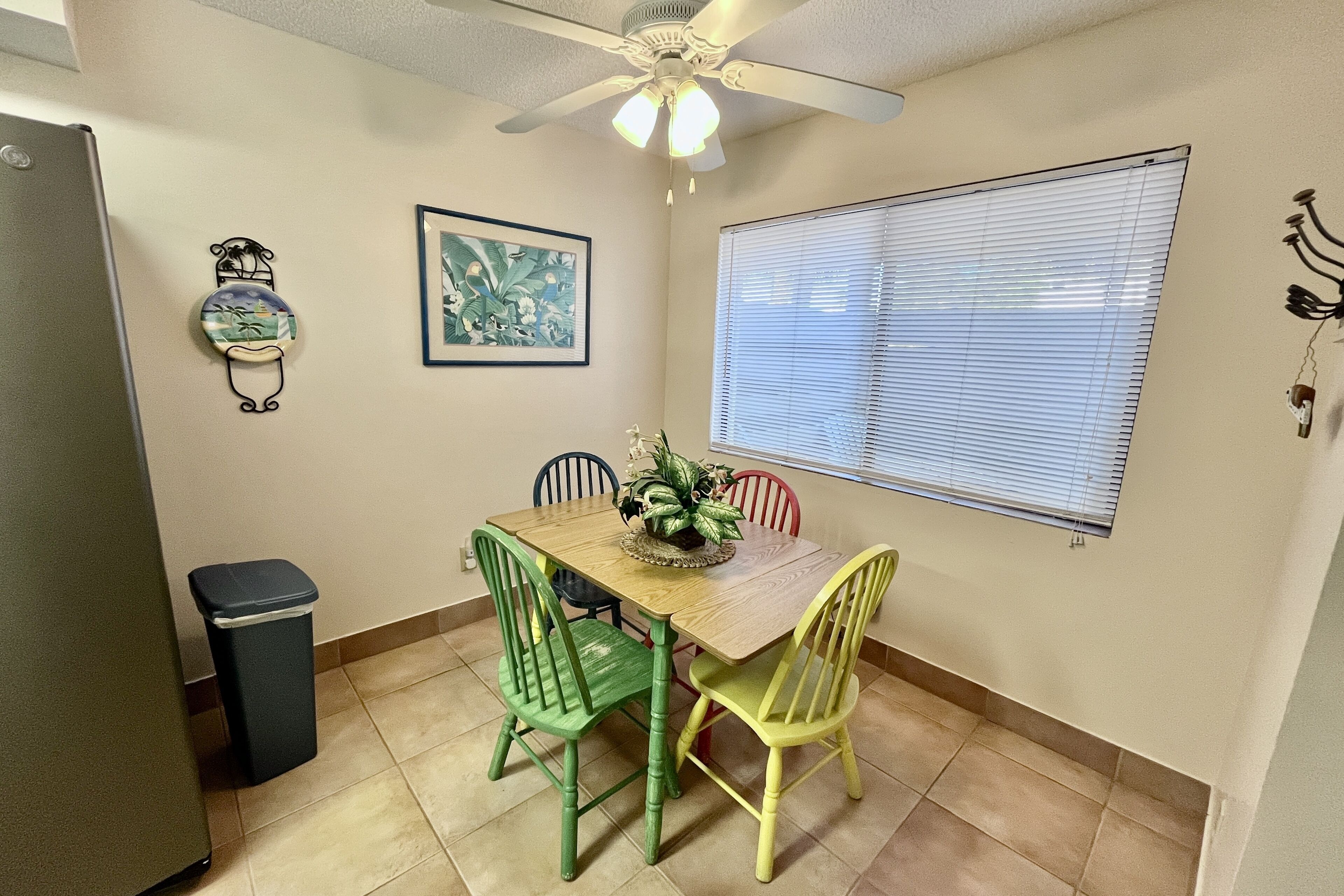 A dining area with a wooden table, four chairs, a ceiling fan, a window with blinds, a trash bin, and wall decorations.